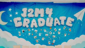 Tường lớp kỷ yếu J2M4 Graduate: chữ bông trắng, trăng lưỡi liềm và ảnh polaroid trên nền xanh đêm