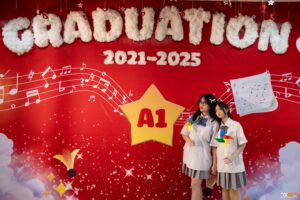 Hai nữ sinh chụp kỷ yếu trước tường vẽ khuông nhạc và huy hiệu lớp A1 niên khóa 2021-2025