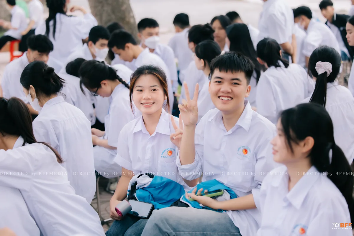 Bạn nữ và bạn nam mặc áo đồng phục trắng cười tươi giữa sân trường đông học sinh