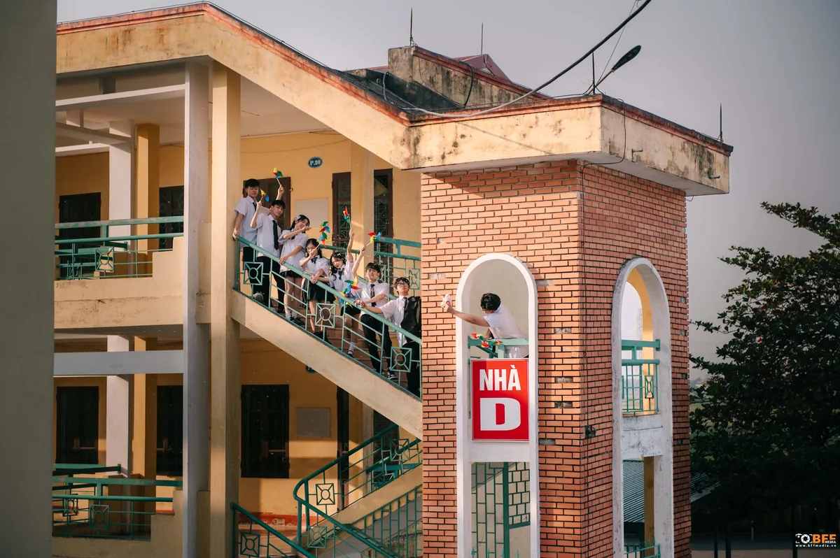 Toà nhà trường có biển Nhà D, học sinh đứng trên cầu thang ngoài trời cầm chong chóng