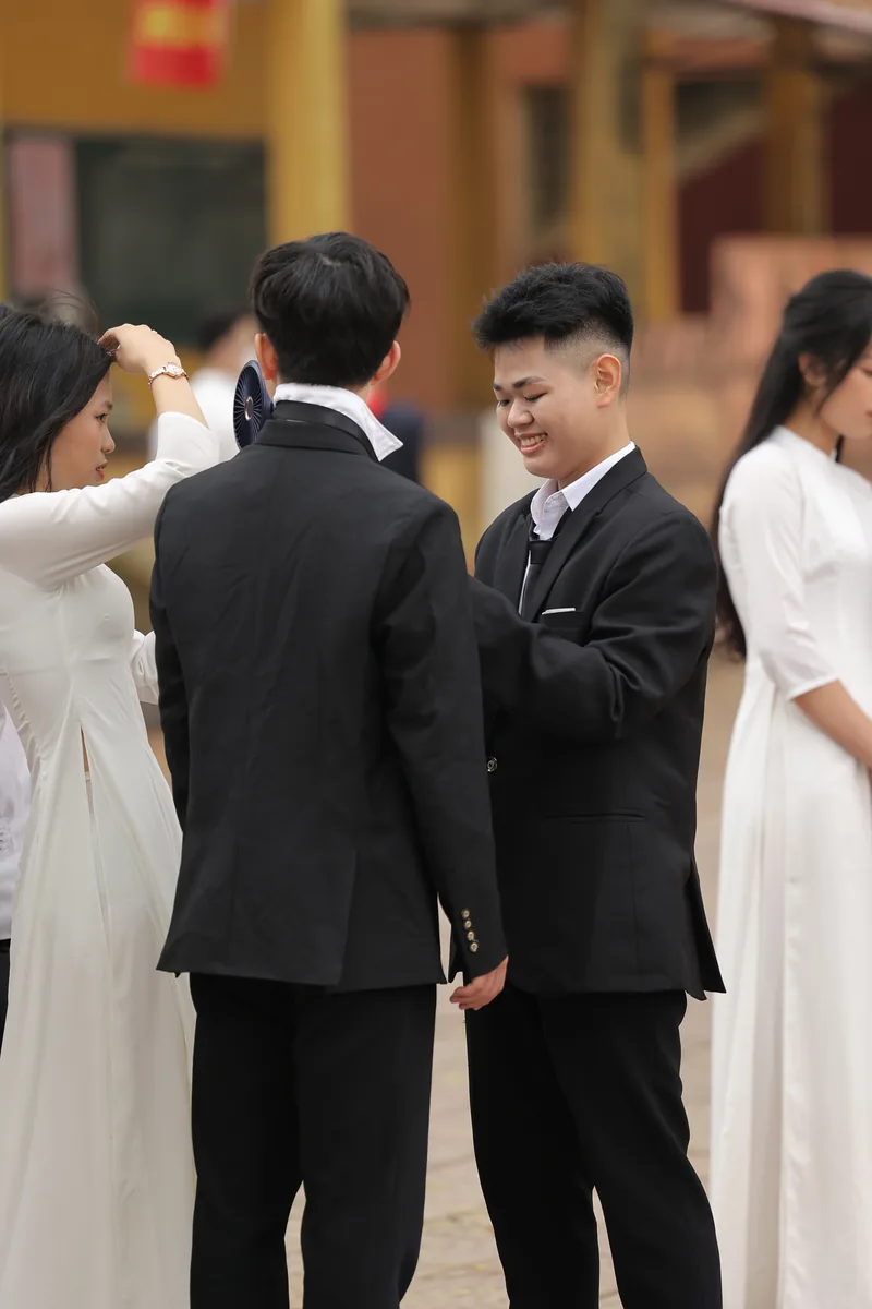 Hai nam sinh mặc vest chỉnh cổ áo cho nhau, bạn nữ áo dài chỉnh tóc cho bạn trước giờ chụp kỷ yếu