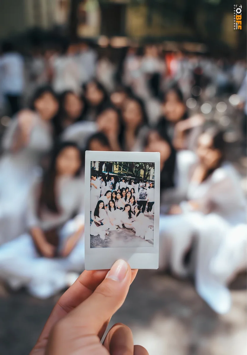 Bàn tay cầm tấm ảnh polaroid chụp cả lớp mặc áo dài trắng ngồi giữa sân trường
